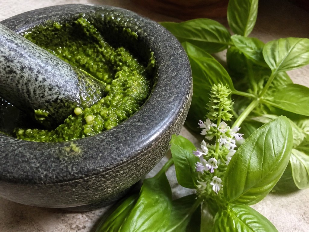 Fresh basil flowers and leaves beside a stone mortar filled with green pesto