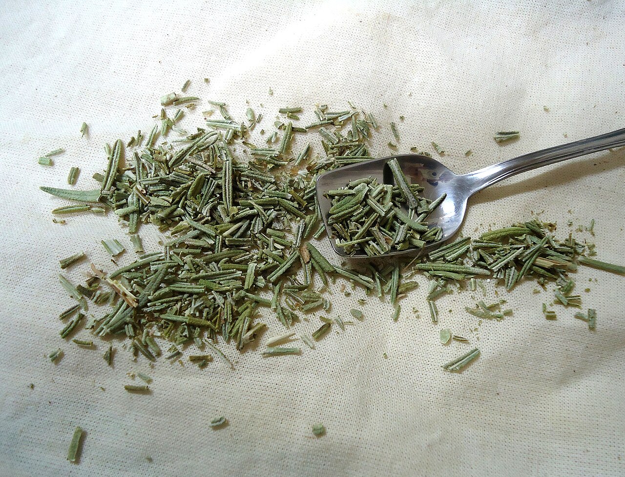 Dried rosemary leaves scattered beside a metal scoop on light fabric